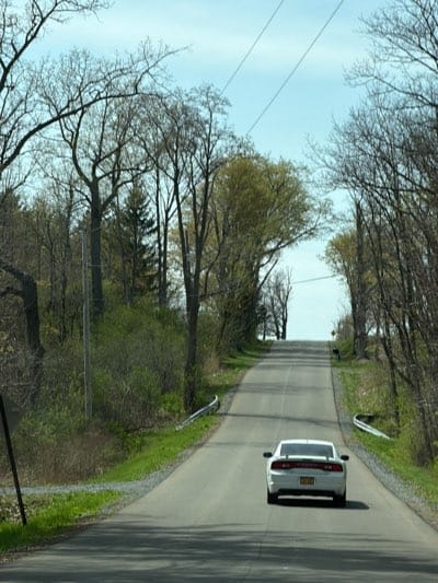 The image captures a view from a road, looking uphill towards the horizon, likely in or near Auburn, United States. A white car, possibly a Dodge Charger, is prominently featured in the foreground, driving away from the viewer up the ascending road. The car's rear, including its taillights and a license plate (illegible), is clearly visible.
The road itself is paved, two-lane, and appears to wind slightly as it goes uphill. Guardrails are visible on both sides of the road at the bottom of the incline. The shoulders are grassy, leading into dense vegetation and trees that line both sides of the road. Many of the trees are deciduous, with bare branches, while others show early signs of spring with nascent green leaves. A few evergreen trees are also interspersed among the deciduous ones.
Above the tree line, the sky is clear and light blue, indicating fair weather. Utility poles and power lines crisscross the upper part of the frame against the sky. Further up the road, a small road sign is discernible on the right side, though its specific message is unreadable. The overall scene suggests a journey through a rural or semi-rural landscape during daytime, with clear visibility. No people are visible in the image.