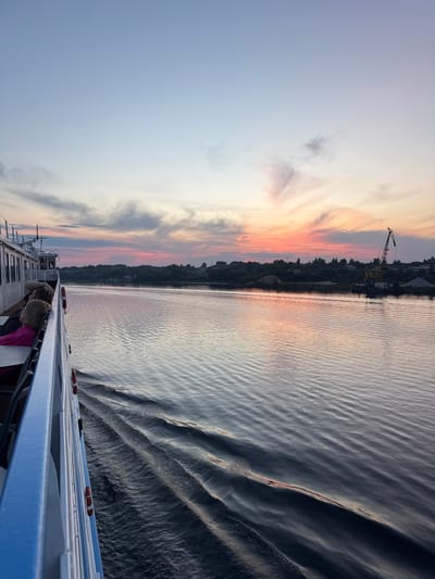 The image captures a view from a boat on the Volga River at sunset, likely near Kimry, Russia. The sky displays a gradient of soft blues and pinks, with wispy clouds tinged by the setting sun. The river's surface is calm, reflecting the sky and showing gentle ripples from the boat's wake.
On the left side of the frame, the white railing of the boat is visible, along with a few passengers seated at tables. One passenger in a pink shirt is prominent. The boat appears to be moving, creating a wake that trails behind it.
The right bank of the river is lined with a dense green treeline. In the distance, a large construction crane stands against the horizon, suggesting an industrial or port area. Piles of sand or gravel are also visible near the shore, indicating some form of material processing or storage. The overall scene conveys a sense of peaceful travel during twilight.
