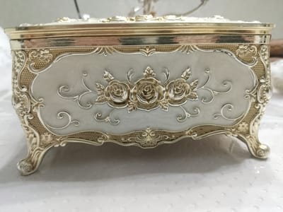This media file features a close-up, eye-level shot of an ornate, decorative rectangular box. The box is a prominent object, presented on a white, textured surface, likely a tablecloth or similar fabric. The background is softly blurred, indicating a shallow depth of field, with hints of a neutral-colored wall or surface.

The box itself is elaborately designed with a combination of metallic gold and a pale, almost pearlescent, light gray or off-white material. The lid and base are bordered by a shiny gold trim. The front of the box is the main focus, featuring a recessed oval panel in the pale material. This panel is adorned with raised, three-dimensional gold detailing. At the center of the panel, three detailed roses are embossed, each surrounded by delicate swirling vines and leaves. The edges of the panel and the surrounding areas of the box are further embellished with intricate gold scrollwork and patterned textures, reminiscent of filigree. The box rests on four ornate, curved legs, also in gold, adding to its regal appearance.

There is no visible text in the image. The lighting is soft and even, suggesting an indoor setting, possibly in daylight or with controlled artificial lighting. The overall impression is one of luxury and classic elegance. The image does not depict any people or specific activities, focusing solely on the aesthetic details of the decorative box. The context of Zhanaozen, Kazakhstan, is not visually apparent within the image itself.