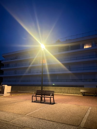 This is a night-time photograph taken at a seaside promenade in Étretat, France. The scene is dominated by a bright street lamp in the center, casting strong rays across the dark blue sky and illuminating a modern, multi-story building in the background. The building appears to be an apartment or hotel, with several balconies and windows, some of which are lit from within. In the foreground, there are two dark-colored benches, positioned side-by-side. The ground is paved with large concrete slabs and a reddish surface. Further to the right, another bench is visible. To the left, a stone monument or plinth is present. The overall atmosphere is quiet and serene, characteristic of a coastal town at night.