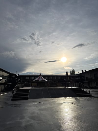 The image shows an exterior scene in Peresvet, Russia. The sky is partially cloudy, with the sun visible. A building with multiple windows and a striped conical structure on the roof is in the mid-ground. A metal stairway and walkway are on the building's side. The foreground appears to be a wet or reflective surface, possibly a paved area.