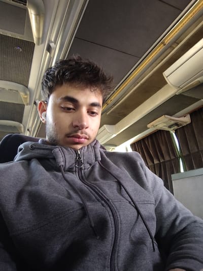 The image displays a young male seated within the interior of a passenger vehicle, consistent with a bus or train, located in the city of Mankat al-Hatab, Syria. The individual possesses dark, textured hair and light facial stubble. He is wearing a grey zippered hooded sweatshirt with visible drawstrings and has a white wireless earbud inserted into his right ear. His head is slightly angled downwards, with his gaze directed off-camera, maintaining a neutral expression. The vehicle's interior features a dark grey ceiling, light-colored overhead panels, and luggage racks. On the right side, a window is partially obscured by brown pleated curtains, below which a small, light-colored rectangular fixture is mounted on the wall.