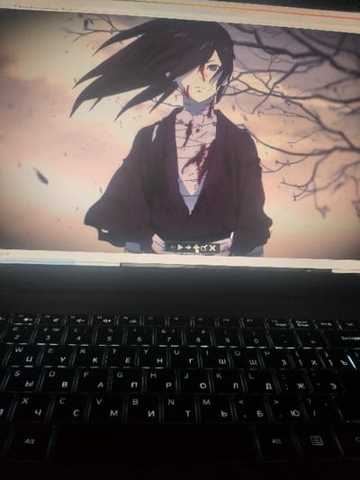 An anime character with long black hair and bandages across their body is displayed on a screen. The character is set against a desaturated background with bare branches and a somewhat bleak, cloudy sky. The laptop screen is illuminated, and the black keyboard is prominently visible to the right. The keyboard's white backlight contrasts with the dark keys, indicating a modern design.