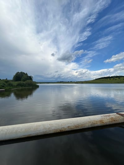 The image depicts a scenic view of a lake in Верх-Рождество, Russia. The foreground is dominated by a weathered, white pipe that stretches horizontally across the lower portion of the frame, partially submerged in the dark water. The lake itself is calm, reflecting the sky and the surrounding landscape. On the left side of the lake, a small island covered in dense green trees and bushes is visible. The distant shoreline features rolling green hills dotted with trees. The sky above is a dynamic mix of bright blue patches and dramatic white and gray clouds, suggesting a variable weather day. No people are present in the image, and no actions or interactions are captured. The overall environment is one of natural beauty and tranquility.