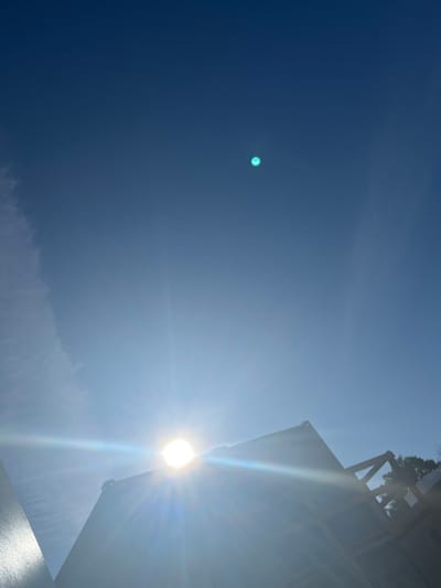 The image shows a clear blue sky with the sun shining brightly. In the lower part of the image, the top of a building structure is visible, with the sun partially obscured by it, creating lens flare effects. The building appears to be made of metal or concrete and has a triangular shape. A faint wispy cloud is present in the upper left portion of the sky. There are no people or animals visible in the image. The location is specified as Jabłonna, Poland.