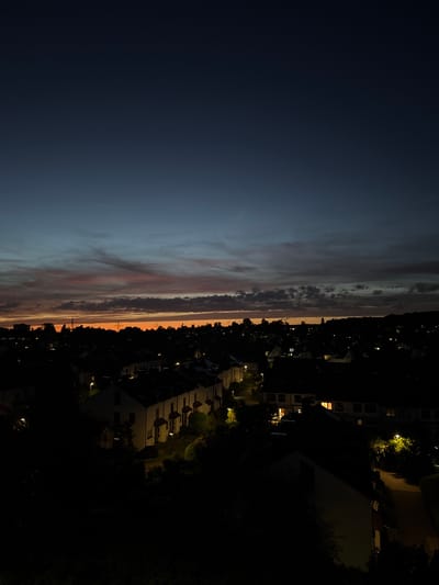 The image depicts a nighttime view of a residential area in Sindelfingen, Germany, during twilight. The sky transitions from a deep blue at the top to an orange and pink hue near the horizon, indicating the end of daylight. Wispy clouds are visible across the sky. Below the sky, a silhouette of trees and buildings forms a dark landscape. The houses appear to be townhouses or row houses, with lights visible in some of the windows, suggesting inhabitants are present. Streetlights illuminate parts of the pathways and gardens between the houses. The overall scene is tranquil and subdued, characteristic of a quiet evening in a suburban setting.