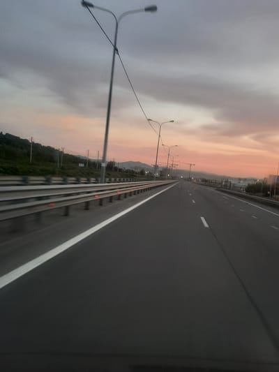 The media file depicts a highway in Mayma, Russia, with a low-angle, slightly blurred perspective suggesting movement. The scene is captured during dusk, as indicated by the soft, warm hues of orange and pink in the sky, contrasting with the darker, overcast clouds. The highway stretches into the distance, with clear lane markings. On the left side of the highway, a grassy embankment is visible, bordered by a metal guardrail. Several streetlights line the highway, with one prominent pole with multiple lights on the left foreground. In the background, distant hills or mountains are silhouetted against the twilight sky. The overall impression is one of a journey or travel during a transitional time of day, with the natural elements of the sky and landscape meeting the man-made structure of the highway. There are no visible people or text in the image.