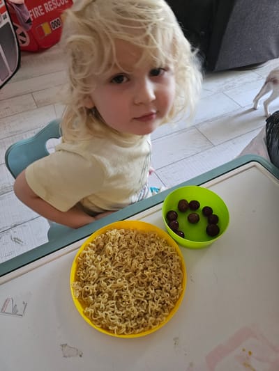 An overhead, eye-level shot captures a young child with blonde hair sitting at a teal plastic high chair. The child is wearing a light yellow t-shirt and is looking directly at the camera with a neutral expression. In front of the child, on a white plastic tray with a green border, are two small bowls. The bowl on the left is yellow and contains cooked instant ramen noodles. The bowl on the right is lime green and holds several dark red grapes. The background consists of a light wood-paneled floor and a glimpse of what appears to be a red toy fire truck. The image is likely taken indoors, possibly in a home. The location is not specified in the image.