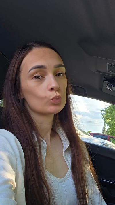In the city of Bulgakovo, Russia, a young woman with long, dark brown hair is captured in a selfie taken inside a car. She is wearing a white collared shirt and a white ribbed tank top underneath. Her lips are pursed in a kiss pose, and she is looking directly at the camera. The interior of the car, including the dashboard and ceiling, is visible. Outside the car, through the window, hints of trees and a cloudy sky can be seen.
