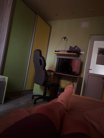 The image depicts an interior scene with a desk, computer setup, and part of a bed. A TV is situated on the desk, with a plush toy of Stitch to its left. Below the desk is a black computer chair. A portion of a bed with pink-toned bedding is visible on the right side of the image, with a person's legs and feet, wearing white socks, resting on the bed. The wall is painted a light yellow-green. There is a window or door visible at the top of the image. The presence of standard European power outlets suggests a location in Europe, and no location-specific items or landmarks are visible to confirm the city of Rakovski, Bulgaria. No specific actions or interactions are visible.