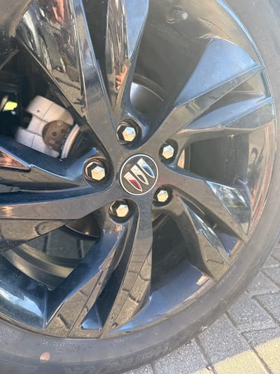 This is a close-up, eye-level shot of a black car wheel and tire, likely taken outdoors on a paved surface. The wheel has a multi-spoke design, and the tire has visible text indicating its specifications. The Buick logo is prominently displayed in the center of the wheel. Some dust and smudges are visible on the wheel's surface. The background shows a textured, light-colored pavement, suggesting an outdoor setting. Sunlight appears to be hitting the wheel, creating reflections and highlights on its glossy surface. The time of day is likely daytime due to the bright lighting. There are no people visible in the frame.
