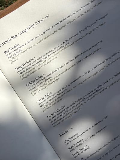 The image shows a close-up of a menu from Atzaró Spa Longevity Juices in Sant Joan de Labritja, Spain. The menu is printed on white paper with black text, and is angled upwards, with a wooden frame visible at the top. The text lists various juices with their ingredients and prices, including "Red Vitality," "Deep Hydration," "Golden Balance," "Green Adapt," and "Matcha Clarity." Below these, there is a section for "Juices 14€" with options like "Mediterráneo," "Mystic Mango," and "Fresh Coconut." Sunlight is casting shadows across the menu. No people are visible in the image.