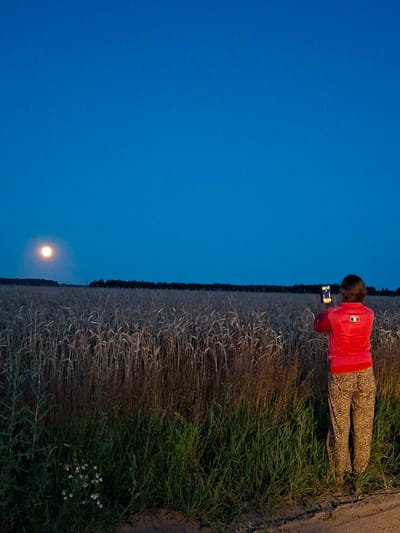 A person wearing a red shirt and leopard-print pants stands in a field of tall, dry grass, facing away from the camera. They are holding a mobile phone up, seemingly taking a picture of the full moon, which is visible in the dark blue sky on the left side of the frame. The moon is a bright, luminous orb, and a faint halo surrounds it. A dark line of trees forms the horizon in the distance. The foreground consists of green grass and weeds, with a dirt path on the right. The scene appears to be set in the evening or early night, with the sky transitioning from dusk to darkness. The location is identified as Slavyenski Sel'ski Sovet, Belarus.