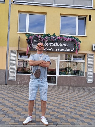 An adult male stands with his arms crossed in front of a yellow building in Kladno, Czechia. The man, appearing in his 40s or 50s, is bald with a light brown, short beard and wears orange-tinted sunglasses. He has a lit cigarette in his mouth, pointed slightly upwards. His attire consists of a light grey short-sleeved t-shirt with a faint logo, light blue distressed denim shorts reaching above his knees, white socks with red and dark blue stripes, and white sneakers with tan soles. A dark brown Nike-branded crossbody bag is slung across his chest. He stands squarely facing the viewer, with his legs slightly apart.

The building behind him is two-storied with a yellow facade. The upper level features two large white-framed windows reflecting a blue sky with clouds. Below these windows, a dark wooden sign, heavily decorated with artificial pink and purple flowers and foliage, displays "Svestkovic" in a script font and "delinárství s kavárnou" (Czech for 'delicatessen with a cafe') in a simpler font. The lower level of the building presents a shopfront with a white-framed glass entrance door on the left and a large display window to its right. The display window contains various items, including a white rabbit figurine, and has decorative patterns applied to the glass. Flanking the shopfront are two distressed white wooden louvered shutters, mounted vertically. An external air conditioning unit is partially visible on the far right of the building, below a small square opening in the wall. The lower portion of the building's facade is textured and colored dark brown-grey. A metal downpipe runs vertically along the left edge of the building.

The ground is paved with rectangular interlocking blocks arranged in a diagonal pattern of alternating light brown/tan and dark grey colors. The scene is illuminated by daylight, suggesting a clear or lightly overcast day.