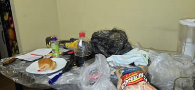 The image depicts a cluttered table surface, possibly in a kitchen or workshop area, covered with a clear plastic sheet. On the table are various items including a partially eaten sandwich on a white plate, a red pen, a blue screwdriver, a yellow power tool, a bottle of dark liquid with a red cap, a black trash bag, a small blue towel with an avocado design, a bag of chips, and several other plastic bags. A tall, clear container with a white lid is visible on the right side of the image. The wall behind the table is painted a light yellow. The overall impression is one of disarray. The image does not contain any specific visual cues that would allow for the determination of its location within Chișinău, Moldova.