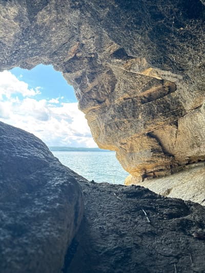 The image shows a view from within a rocky cave looking out towards a body of water and a cloudy blue sky. The foreground is dominated by the dark, rough texture of rocks, with a sliver of the open water and sky visible through the cave's opening. The rocks forming the cave mouth are layered and show variations in color, from dark grey to lighter brown and tan hues, suggesting a natural geological formation. The water appears calm with small ripples, and a distant shoreline with trees is faintly visible under the bright sky. There are no people or animals visible in the scene. The location is identified as Dom Otdykha Zolotoy Plyazh, Russia.