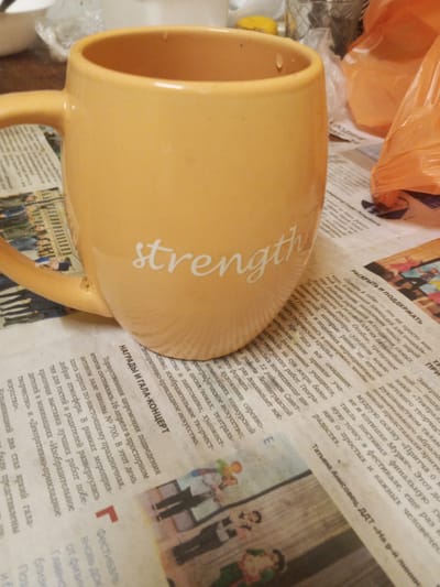An overhead shot shows an orange mug with the word "strength" printed on it, resting on a newspaper. The mug's handle is positioned to the left of the frame, and it is partially filled with an unseen liquid. The newspaper contains text in Russian and images of people, likely from a local event or publication. The background is out of focus, revealing what appears to be a table with other objects, including a plastic bag. The scene is dimly lit, suggesting an indoor setting, possibly a home or office. There are no people visible in the frame, and the mug is the primary subject. The newspaper headline appears to be in Russian. Without further information or context, it is impossible to determine the specific location of Ulyanovka, Russia.