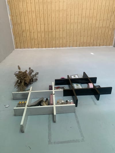 The image depicts an interior scene with a light blue, concrete floor and a light-colored wall in the background that transitions into a wood-paneled wall at the top. In the foreground, there are two white shelves and one black shelf unit.
The white shelf unit on the left is L-shaped and has several compartments. It holds a bouquet of dried flowers in a glass container, various small bottles with golden caps, a stack of papers, and a pink cylindrical object.
The black shelf unit is a tic-tac-toe design and is placed to the right of the white shelf. It contains two framed certificates or documents with green emblems, a pink roll of tape, and a black object that appears to be an electronic device.
The lighting in the scene is diffuse, with some reflections visible on the floor. The overall impression is of an organized display of items on shelving units in what appears to be a room under renovation or in a state of disuse. There are no people visible in the image. The location is identified as Meunasahlhok, Indonesia.