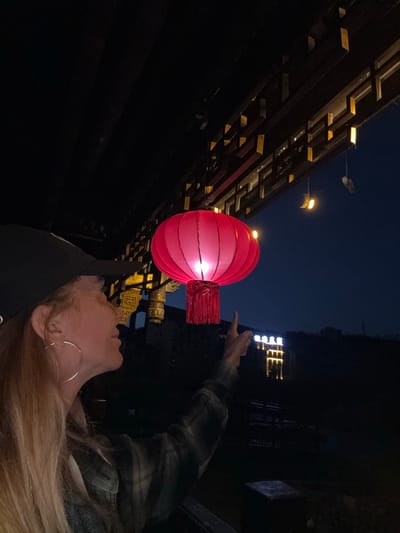 At night, in what appears to be Zhuoshui, China, a person is pointing up towards a glowing red lantern. The environment includes architectural features with traditional Chinese design elements, such as intricate woodwork. Streetlights are visible in the background. The person in the photo has blonde hair and is wearing a denim jacket over what appears to be a black shirt. A large hoop earring is visible.
