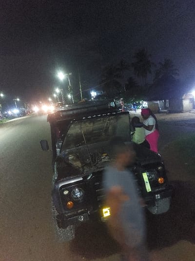 The image depicts a nighttime scene in Msata, Tanzania. A dark-colored off-road vehicle, resembling a Land Rover Defender, is parked on the unpaved shoulder of a paved road, with its hood open. On the front passenger side of the vehicle, a woman wearing a white top, pink lower garment, and a pink head covering stands alongside a child in a light green top, who appears to be held or positioned close to her. A third individual, heavily blurred by motion, is partially visible in the immediate foreground, wearing a light-colored shirt and facing away from the camera. The paved road stretches into the distance, illuminated by multiple streetlights and the headlights of other vehicles. To the right of the scene, several low-rise structures, possibly buildings or market stalls, are visible, illuminated by artificial light, with palm trees silhouetted against the dark sky. The open hood of the vehicle suggests a mechanical inspection or breakdown.