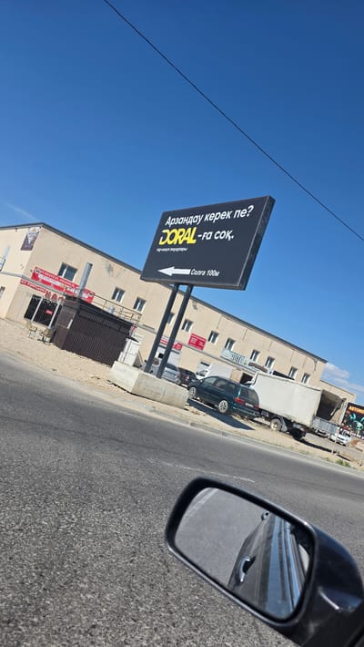 The image depicts a street view in Баянды ауылдық округі, Kazakhstan, on a clear sunny day. In the foreground, a car's side mirror reflects the road and the car's side. The middle ground shows a paved road with a sidewalk and a row of buildings. A large black billboard with yellow and white text is prominently displayed, advertising "DORAL -fa соқ." and indicating "Солға 100м" (Left 100m). The buildings appear to be commercial, with signage visible, including "TURKESTAN SHASHLIK AL" and what looks like a loading dock. Several vehicles are parked along the street, including a dark green sedan, a white truck, and a white car further down. The background is dominated by a bright blue sky with a single black wire stretching across it. The overall scene suggests a typical roadside commercial area in a rural or semi-urban setting in Kazakhstan.