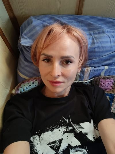 The media file is a close-up, high-angle selfie of a woman lying down. She has short, pinkish-blonde hair and is wearing makeup, including eyeliner and mascara. Her lips are painted a light pink. She is wearing a black t-shirt with a white graphic print that appears to be an arm making a rock and roll hand gesture. She is lying on a bed with a blue striped pillow and a colorful crocheted blanket underneath her. The background shows a wall with vertical wooden slats, suggesting an indoor setting. The lighting appears to be natural, possibly from a window, and indicates daytime. The woman's expression is neutral. There is no visible text in the image. The location context is given as Maurino, Russia.
