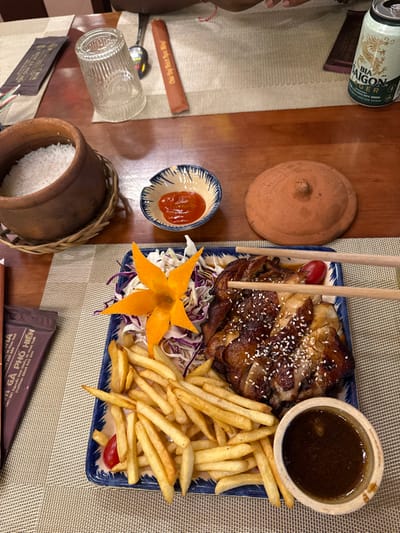 This image depicts a meal served on a wooden table within a restaurant located in Thành phố Phan Thiết, Vietnam, identified by a "GÀ PHỐ HIỆN" packet displaying the address "341 Hùng Vương, P.Phú Thủy, TP.Phan Thiết." The central subject is a rectangular plate holding grilled chicken, garnished with white sesame seeds, accompanied by a large portion of french fries, a small shredded cabbage salad, a star-shaped carrot carving, and two cherry tomatoes. A pair of light-colored wooden chopsticks is positioned over a piece of chicken. Adjacent to the plate are a small terracotta bowl containing dark dipping sauce and a small ceramic bowl with red chili sauce. To the left, a terracotta pot of white rice rests on a woven coaster. Other items on the table include an inverted clear glass, a silver spoon, a brown chopstick wrapper, a round terracotta lid, and a partially visible can of Bia Saigon Lager beer. A person's arm is visible at the top, indicating an active dining scene.