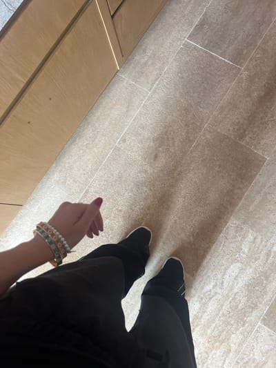 A high-angle shot shows a person wearing black pants and black sneakers standing on a light brown tiled floor. A wooden cabinet is visible in the lower left corner of the frame. The person's left hand is in view, adorned with multiple bracelets. The background is a light brown tiled floor.