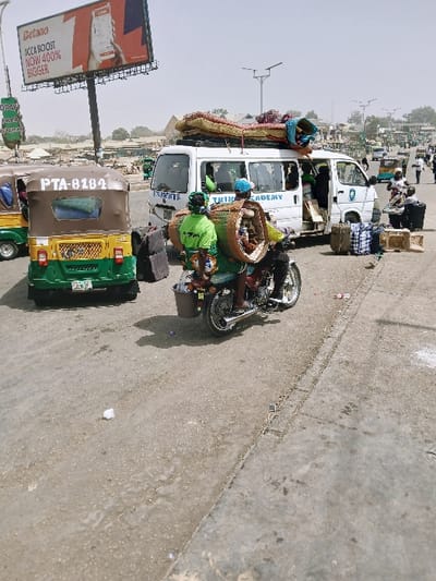 The image depicts a busy street scene in Masvingo, Zimbabwe, under a hazy sky. In the foreground, a motorcycle is in motion, moving left across the frame. It carries a driver wearing a blue cap and a passenger in a green shirt, who is transporting a large cylindrical woven basket on their back and a smaller bucket attached to the motorcycle's side. Adjacent to the motorcycle on the right, a white minibus, marked "STUDENTS" and "TRINITY ACADEMY," is stopped with its side door open. The minibus is heavily loaded with cargo, including mattresses and bundled items on its roof rack. Multiple suitcases, duffel bags, and boxes are positioned on the ground beside its open door, indicating loading or unloading activity. To the left of the minibus, a green and brown autorickshaw, bearing the license plate "PTA-0104," is oriented facing away from the viewer. A large billboard advertising "Betano" with "ACCA BOOST NOW 400% BIGGER" is visible high on the left. The asphalt road surface exhibits wear and scattered debris. In the background, additional pedestrians, vehicles, and low-rise buildings are present, along with multiple street light poles.