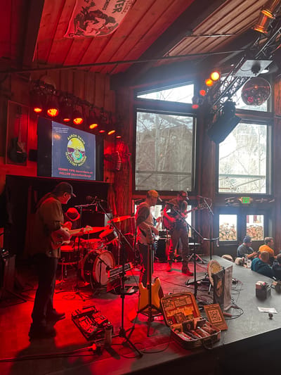 A live band performs on stage in a rustic venue with wood paneling. They are illuminated by red stage lights. A disco ball hangs overhead, and large windows reveal an outdoor scene. A screen displays the band's name, "Aaron Davis & The Mystery Machine," along with their Venmo tip information and social media handle. A poster hangs on the wall nearby. Patrons are seated at a bar in the background.