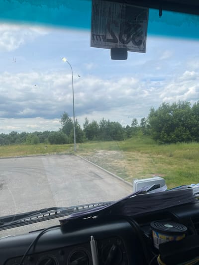 The image, taken from inside a vehicle's cabin, captures a view through the windshield. The windshield is moderately dirty with smudges and what appear to be small specks of debris. In the upper section of the windshield, a blue tint is visible, likely from a sun visor or tinting. A sticker, partially obscuring the view, is affixed to the windshield.
Outside, the scene depicts a cloudy sky with scattered white clouds. A streetlamp with an illuminated bulb stands to the left of the frame. In the middle ground, there is a paved surface, possibly a road or parking area, which transitions into a grassy field with scattered trees and bushes forming a treeline in the background. The environment appears to be a semi-rural or outskirts area, given the presence of natural vegetation and limited urban infrastructure.
The foreground shows the interior of the vehicle, including a dashboard with various controls and a stack of papers. A small, black air freshener is also visible on the dashboard. No people are discernible in the image. The overall impression is a still shot of a location from a stationary vehicle, likely during daytime.