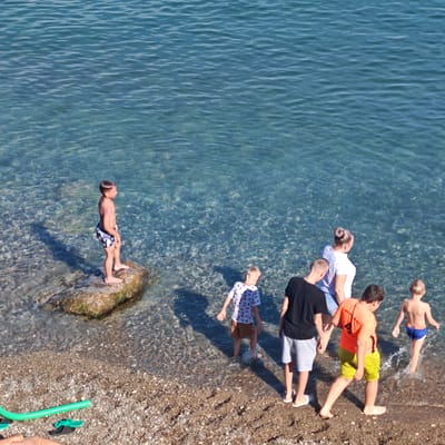 This media file captures a sunny day at a pebble beach in Simeiz, Ukraine. The clear, shallow water of the Black Sea laps at the shore, revealing the pebbly seabed. Several people, including children and adults, are enjoying the seaside.
On a large rock near the water's edge, a young boy in black and white patterned swim trunks stands looking out at the sea. A group of people are in the shallow water and on the wet sand. Among them is a young boy in a white t-shirt with a patterned design and brown shorts, his back to the camera. Next to him, a boy in a black t-shirt and grey shorts stands. Further to the right, a boy wearing an orange t-shirt and yellow shorts walks into the water. Another young boy, wearing blue swim trunks, is already wading in the water. An adult woman in a white t-shirt and blue jeans is also present.
A bright green tube lies on the shore, along with a pair of black and green flip-flops. The sunlight casts distinct shadows on the water and the beach, suggesting it is daytime, likely mid-morning or afternoon. The overall mood appears relaxed and recreational, typical of a beach outing. No visible text is present in the image.