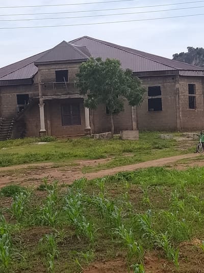 A low-angle, medium shot captures a large, unfinished, two-story house with a metal roof in Barikin Sibre, Nigeria. The building's exterior walls are constructed from grey cinder blocks, and several windows are open, revealing dark interiors. A set of concrete stairs leads to an upper-level entrance, flanked by a white pillar. A large tree stands in front of the house, and a person is visible on the right side of the frame, riding a bicycle. In the foreground, young corn plants grow in a field. The sky above is overcast, and power lines are visible stretching across the frame. In the background, a rocky outcrop rises above the treeline.