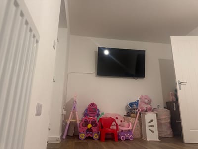 A photograph taken indoors depicts a room with various objects, most notably children's toys. The room features white walls and a dark wood floor. On the wall hangs a flat-screen television. In front of the wall, a collection of toys is arranged including a pink play vanity, a red plastic chair, a purple walker toy, and a pink teddy bear. A white storage unit stands to the right of the toys. In the corner, near a white door, there's a cabinet with a couple of stuffed animals atop it. The setting appears to be a playroom or child's bedroom. The location is the Vale of White Horse, United Kingdom. No people are visible in the photograph.