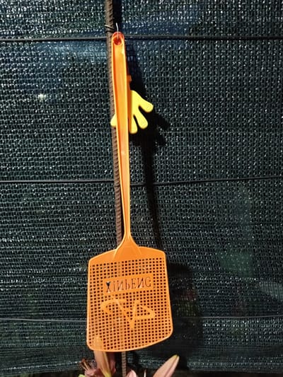 An orange flyswatter hangs on a rebar against a dark green mesh fence in Shchastia, Ukraine. The flyswatter has a yellow hand-shaped accessory attached to its handle. The words "Хибень" are visible on the flyswatter's perforated surface. In the lower part of the image, the tops of some plants are visible. The lighting suggests it may be dusk or dawn.