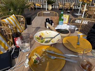 The image depicts an outdoor dining area on a wooden deck, identifiable as a restaurant or beach club in Los Alcázares, Spain, marked by a "3 PELAOS" QR code stand. The scene is illuminated by direct sunlight, casting distinct shadows.
In the foreground, a light-colored wooden table is set with used dishes and various items. A large white bowl, centrally located, contains remnants of a salad, with two spoons resting inside. To its immediate right, a mustard-yellow oval plate is largely empty, with a single fork. To the left, another similar mustard-yellow oval plate holds residual salad and a fork and knife.
Beverage containers include a green S.PELLEGRINO sparkling water bottle, two clear ribbed glasses (one containing water, one empty), and a tumbler with a dark liquid and ice cube. Condiments on the table consist of a dark glass bottle of ARMONIA olive oil and a tall clear shaker containing pink salt. A small dark wooden bowl with a white napkin and a black rectangular holder are also present. Personal items such as a dark wallet or phone, a set of keys, and a white charging cable are visible on the table. A person's leg, clad in dark trousers, is partially visible on the far right edge of the frame.
The background shows additional wicker chairs with yellow and white striped cushions arranged around tables on the wooden deck. Palm fronds extend from the left, and potted plants in yellow and white striped vases are positioned throughout the background, with parts of light-colored outdoor umbrellas also visible.