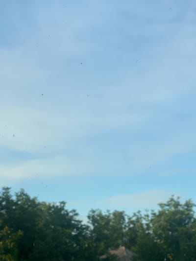 The image shows a blurry view of a blue sky with some wispy clouds. In the foreground, there is a line of green trees that are also out of focus. The bottom of the image also shows a hint of a light-colored roof, possibly belonging to a building. The overall impression is that the photo was taken from a window, looking out at a natural scene on a clear day. There is no visible indication of the location in Vălcineț, Moldova.