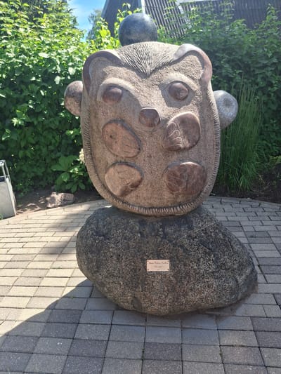 An outdoor stone sculpture, resembling a stylized hedgehog or similar creature, is positioned on a paved area in front of lush green bushes. The sculpture is large and textured, with carved facial features and round, lighter-colored stone inlays that suggest spots or paws. A smaller, rounded stone rests on top of the sculpture's head, and two smaller, spherical stone elements protrude from its sides, perhaps representing ears or cheeks. The sculpture sits atop a much larger, rougher boulder, to which a small, rectangular plaque is affixed. The plaque has text and appears to be made of brass or a similar metal. The paved area is made of interlocking rectangular stones. The overall scene suggests a public art installation or a garden feature. The plaque has been identified as bearing the inscription "Agnese Rutkaite-Kirillova. "Gudra maza". 2014." This sculpture is located in the city of Ogre, Latvia, in the central park area along the river.