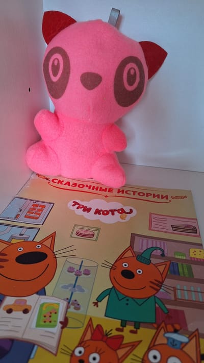 A pink plush toy, possibly a panda, is positioned in the foreground, leaning on a children's book. The book is open, revealing colorful illustrations of cartoon cats and a kitchen scene. The text on the book appears to be in Russian. The background is a plain white surface, likely a shelf or wall. The lighting is bright and even, suggesting an indoor setting. There are no visible people or actions in the image. The image does not contain any specific landmarks or elements that would allow for the determination of its location in Nogliki, Russia.