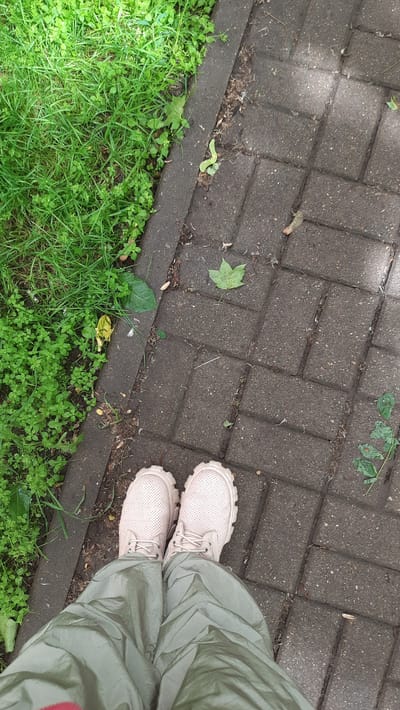A pair of light pink perforated lace-up shoes are worn by an unseen person standing on a paved walkway made of grey interlocking rectangular bricks. The walkway is bordered by a strip of green grass and small plants. Several fallen leaves, one prominent green leaf, and some debris are scattered on the bricks. The person is wearing loose-fitting, olive-green pants. The image captures a close-up perspective of the lower body from the knees down. The scene appears to be outdoors in a park or a paved urban area.