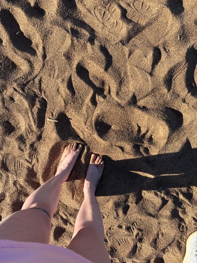 An overhead, full shot captures a person's legs and feet standing on a sandy beach. The sand is coarse and uneven, with distinct footprints and shadows cast by an unseen light source, likely the sun, indicating it is daytime. A thin, dark band tattoo encircles the left thigh. To the far right edge of the frame, a small portion of a white sneaker with blue detailing is visible. The scene is devoid of any other individuals or notable objects, emphasizing the focus on the person's lower extremities and the beach environment. The location is Zelenogorsk, Russia.