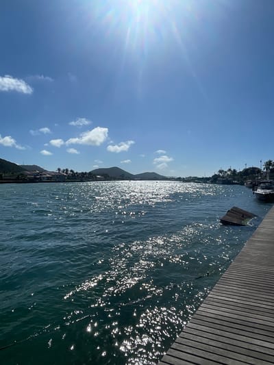 The image depicts a sunlit coastal scene. The perspective is from a wooden boardwalk or dock, with the boards running along the right side of the image. Water, reflecting intense sunlight, dominates the central and right portions. Across the water, the landscape includes a coastline with trees and buildings, possibly a residential area. Distant hills or mountains are visible. The sky is blue with scattered, puffy white clouds and a bright sun in the upper left corner. Given the prompt information, the location is likely Cabo Frio, Brazil.