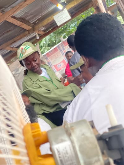 In the city of Orun Ekiti, Nigeria, an outdoor scene unfolds under a makeshift corrugated metal roof supported by wooden beams. A young woman, wearing a green uniform and a matching cap adorned with a national emblem, is the central figure. She is looking down, possibly in conversation or deep in thought, with her hair pulled back and a few strands falling forward. To her right, the back of another person's head is visible, also with dark, curly hair.

The background reveals a group of individuals, including a person in a pink top and another in a grey shirt, partially obscured. Stacks of what appear to be cement bags or similar materials are visible behind them, some bearing red and white markings. The environment seems to be an informal gathering space, possibly a temporary shelter or a market area.

In the foreground, a white electric fan, partially disassembled, is prominent. Its yellow plastic components and metallic parts are in focus, suggesting it might be undergoing repair or is simply present in the scene. The overall lighting is natural, hinting at daylight hours.