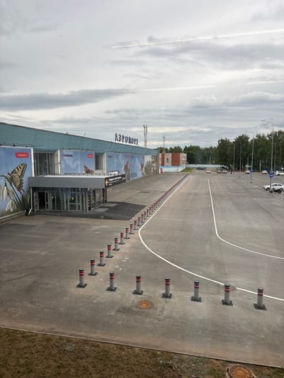 This media file shows an exterior view of an airport building, likely in Старое Мартьяново, Russia. The building has a light blue facade with large billboards featuring images of butterflies and text that appears to be in Russian. One billboard clearly shows the word "ИЖАВИА". Above the billboards, the word "АЗРОПОРТ" is visible, which translates to "Airport". The building has glass doors and windows, suggesting an entrance or reception area.

In the foreground, there is an asphalt area with white markings, indicating a road or parking area. Several cylindrical bollards with red and white stripes are lined up along the asphalt. To the right of the asphalt, there is a strip of dry grass and a circular manhole cover. In the background, there is a line of trees and a cloudy sky. A few cars are parked in the distance, and a white motorcycle is visible near some of the cars. The overall scene suggests a daytime setting with overcast weather. There are no people prominently visible in the image, and the focus is on the architectural elements and surrounding environment of the airport.
