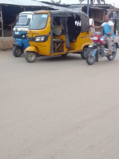 The image shows a yellow three-wheeled auto-rickshaw parked on the side of a paved road in Adanisievbe, Nigeria. The rickshaw is parked beside a smaller, blue auto-rickshaw and in front of a blue motorcycle. A man wearing a blue shirt is riding the motorcycle. The scene takes place on a street in a residential area. The sky is overcast.
