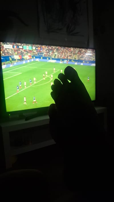 The image is a close-up shot of a television screen displaying a soccer match. The score on the screen shows France 0 and Spain 1, with the game time at 53:44. The television is turned on and emitting light, illuminating the dark room. In the foreground, a silhouetted human foot is visible, obscuring part of the television screen. The foot appears to be resting or placed in front of the television. The background is dark, suggesting the room is dimly lit or the flash was not used. The overall scene conveys a relaxed atmosphere of watching television, likely in a home environment. There is no specific indication of the location being in the city of Çiğli, Turkey, beyond the provided context.