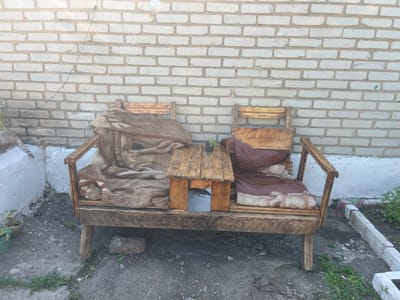 A wide shot of a rustic wooden bench with two armrests and a small wooden table between them, placed against a light-colored brick wall. The bench is covered with two blankets: a patterned brown and beige blanket on the left side, and a reddish-brown blanket on the right side. A small potted plant sits on the table. The ground in front of the bench is rough asphalt with some weeds and debris. To the left of the bench, a portion of a white wall and a green potted plant are visible. A cat's tail is also visible on the white wall. To the right of the bench, a concrete curb with some greenery is visible. The overall scene appears to be an outdoor seating area. The location is identified as Oktyabrskoye, Kazakhstan.