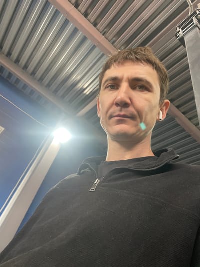 A low-angle, close-up, selfie-style shot features a man with short brown hair and light green eyes looking directly at the camera. He is wearing a black zippered jacket and a white wireless earbud in his right ear. A light blue rectangular reflection is visible on his left cheek. The background consists of a corrugated metal ceiling, a bright light fixture, and a portion of a blue wall with a white trim. The scene appears to be indoors, possibly in a warehouse or industrial building, with no visible indications of the city of Сокуры, Russia.