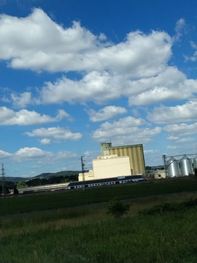 The image depicts an industrial area with several large grain silos and storage facilities, set against a backdrop of rolling green hills under a bright blue sky dotted with white cumulus clouds. A tall, yellow and green building with multiple windows stands prominently, possibly a grain elevator or processing plant. To its right, a row of shiny, silver silos rises, reflecting the sunlight. A lower, cream-colored building with numerous windows extends from the base of the yellow structure. Electrical pylons and power lines are visible throughout the scene, indicating the presence of industrial infrastructure. The foreground features a grassy field with a dark green slope leading up to the industrial buildings. The location is identified as Kapsova Lhota, Czechia. There are no people visible in the image.