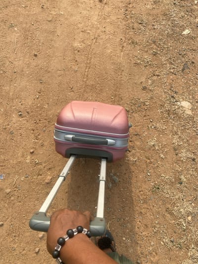 An overhead, slightly angled view depicts a person's right arm and hand pulling a rose-gold, hardshell wheeled suitcase across an unpaved, reddish-brown dirt path in Odo Ona, Nigeria. The hand, adorned with a bracelet of dark spherical beads interspersed with clear rectangular beads, grips the gray handle of the fully extended, silver-colored telescopic pull rod of the suitcase. The suitcase features a textured exterior and a gray circumferential accent stripe. The ground is composed of compacted earth, scattered with small stones, pebbles, and patches of dry, light-colored vegetation. Distinct tire tracks are impressed into the dirt surface. Part of the person's right foot, wearing a dark sandal, is visible behind the pulling hand, along with a portion of green fabric, likely trousers.